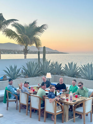 Fish Ranch Hosted Group in La Ventana enjoying a dinner on the patio overlooking a ocean view at sunset, with palm trees, agave plants, and mountains in the background.